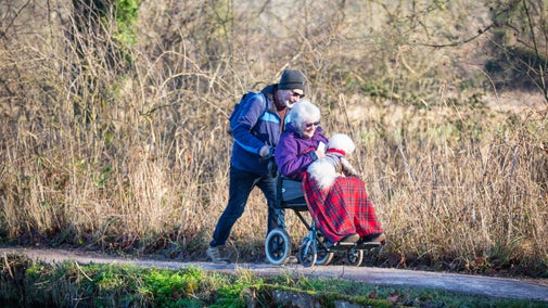 Two people, one of whom is in a wheelchair and has a dog in their lap, are walking alongside the riverside in winter at Mottisfont, Hampshire.